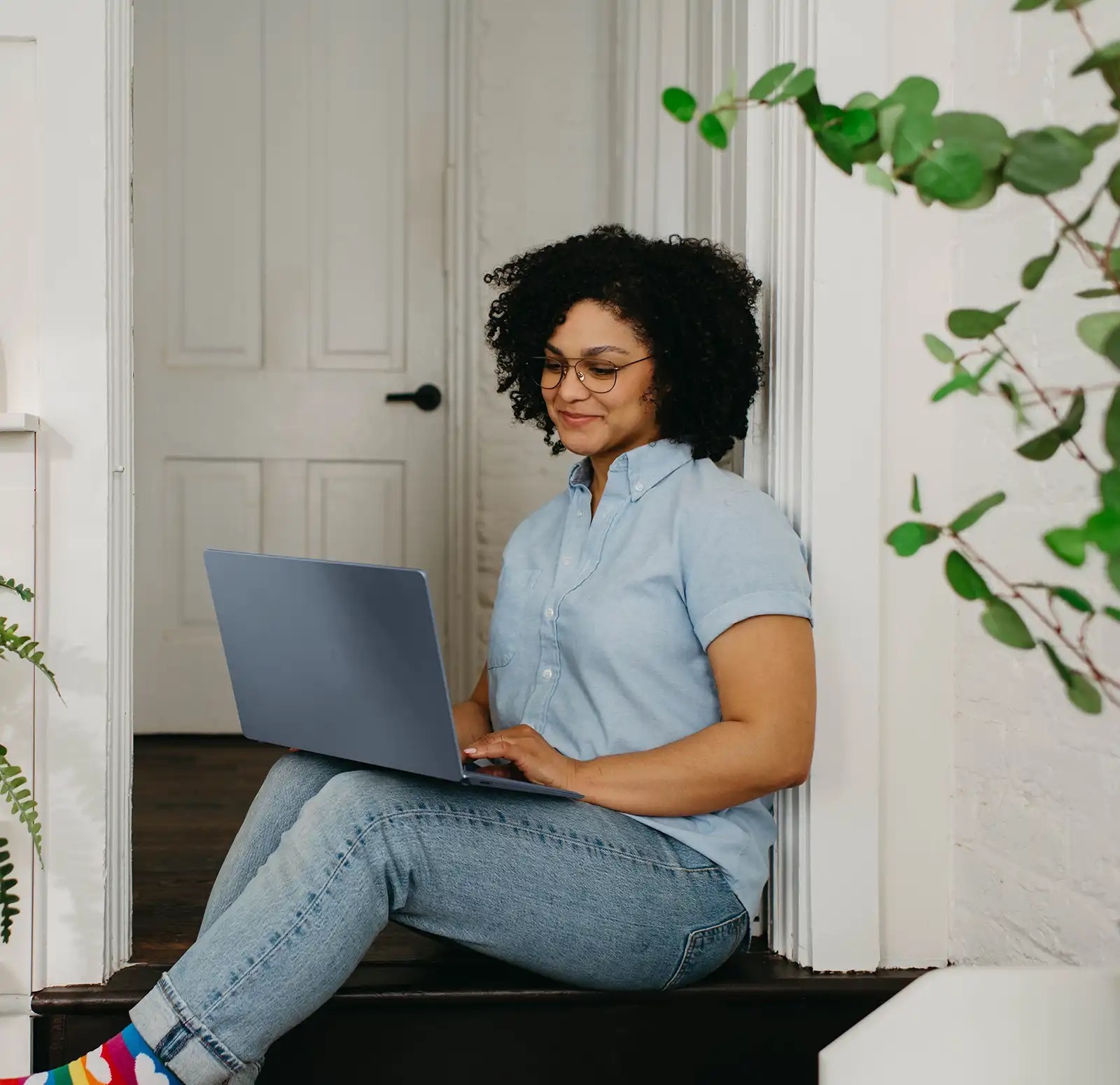 A curly haired woman sitting casually on her doorstep, working on a laptop.