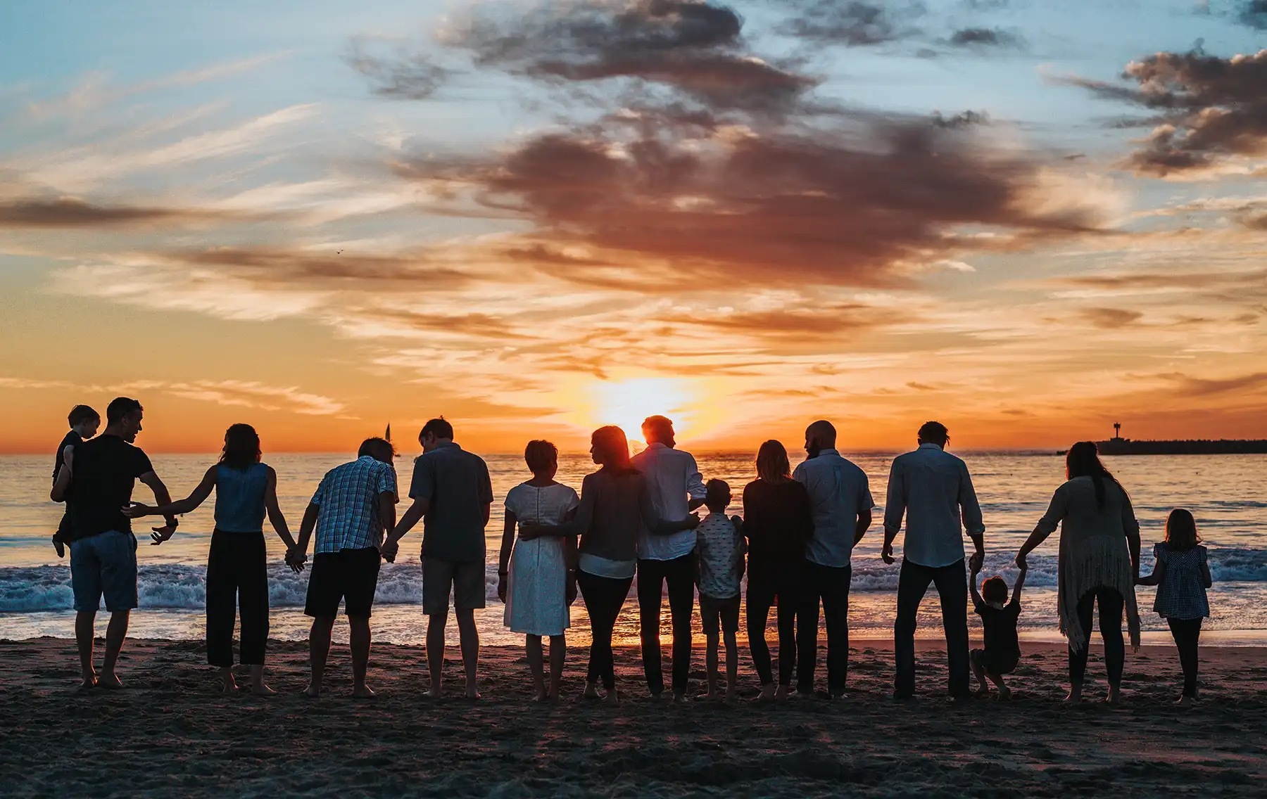 Large, multi-generation family holding hands on the shore of a beach, silhouetted by the sunset.
